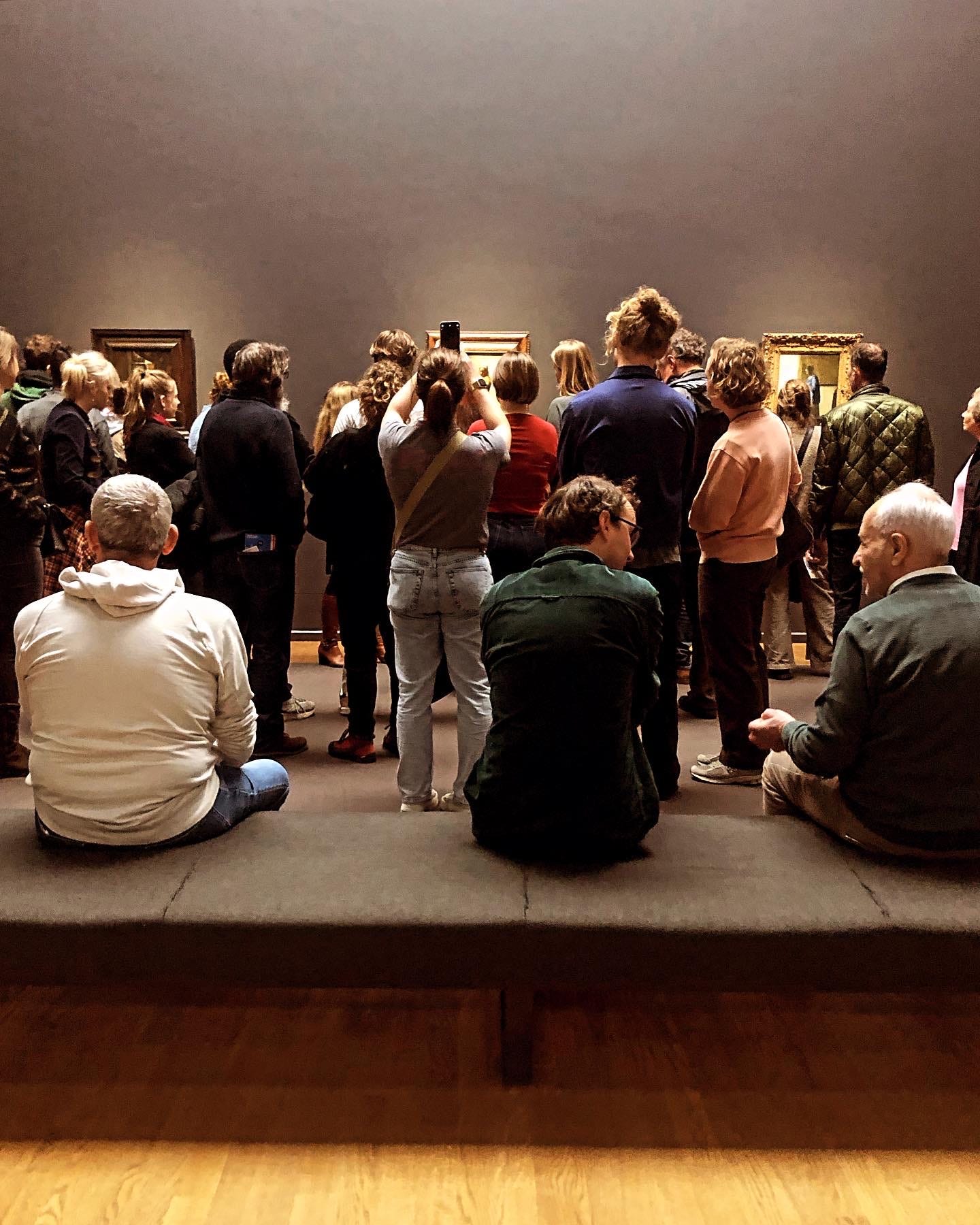 A large group of people stand looking at Vermeer's 'The Milkmaid', almost entirely obscuring the camera's view. One woman holds a cameraphone aloft. In the foreground, three men sit on a low bench. Two of them are in conversation. The room feels busy, but hushed. Almost everyone's attention is angled in towards the Vermeer painting, which looks very small from here.