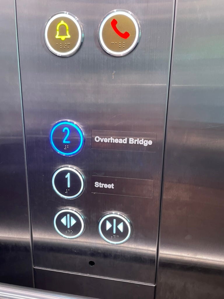 Lift button panel with large buttons. Level 2, Overhead bridge, is active and glowing blue. Below are level 1, street, and open and close buttons. Above are the bell button and the call button. Each button is equipped with Braile.