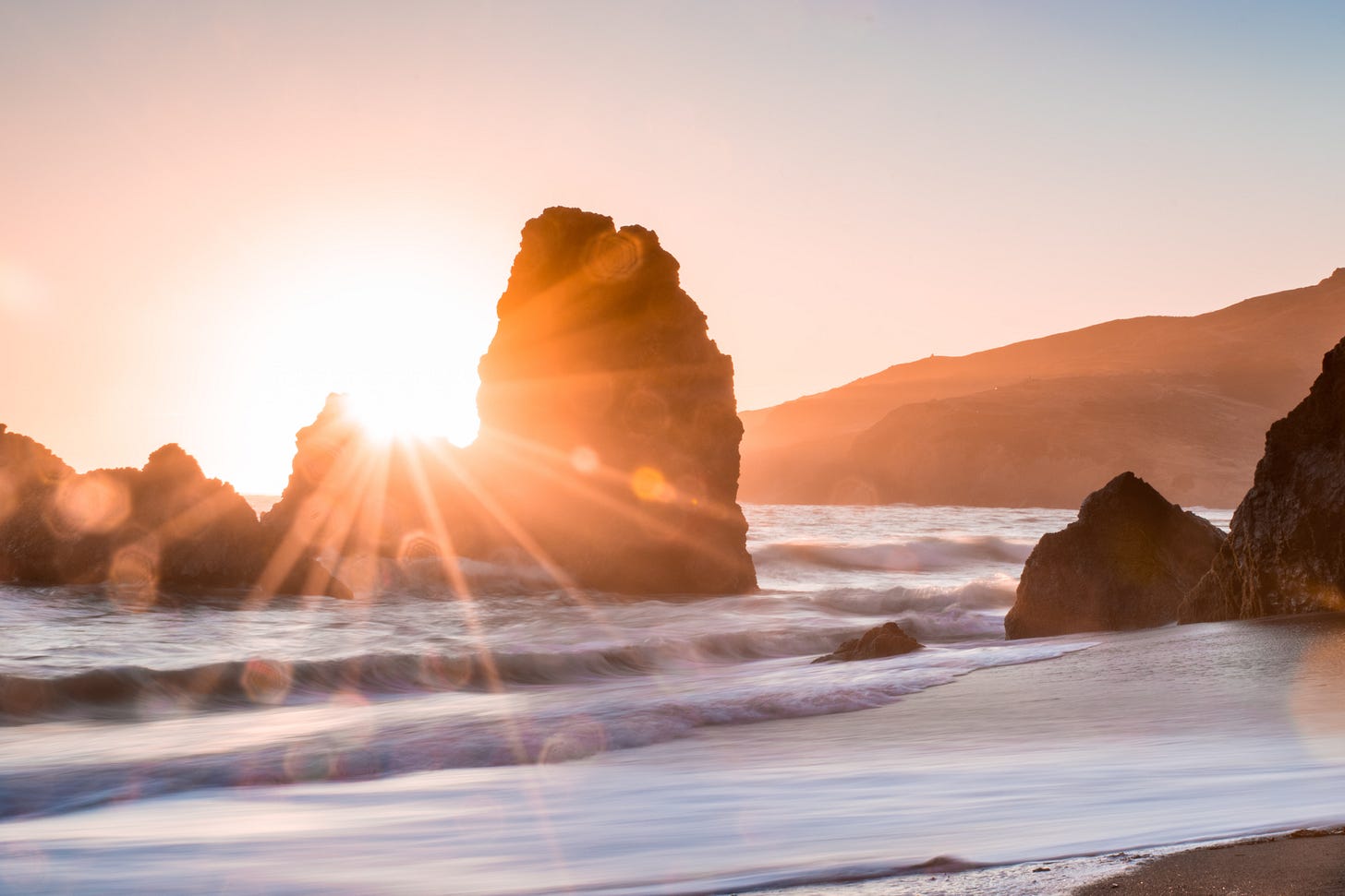 Sunrise in between rocks. The rocks are surrounded by water, with coastline in the foreground.