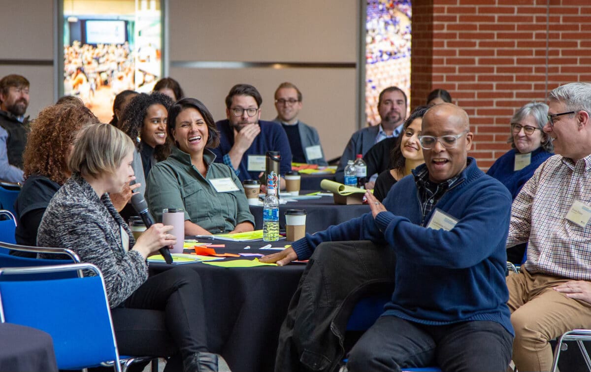 A wide-angle, horizontal photo of a professional media summit. A group of diverse people are seated around a circular table covered in a black cloth with yellow notebooks and coffee cups. In the foreground, a woman with blonde hair speaks into a microphone while others in the background—including a woman in a green jacket and a man in a blue sweater—laugh and engage in the conversation. The setting is a brightly lit indoor space with large windows and a brick pillar.