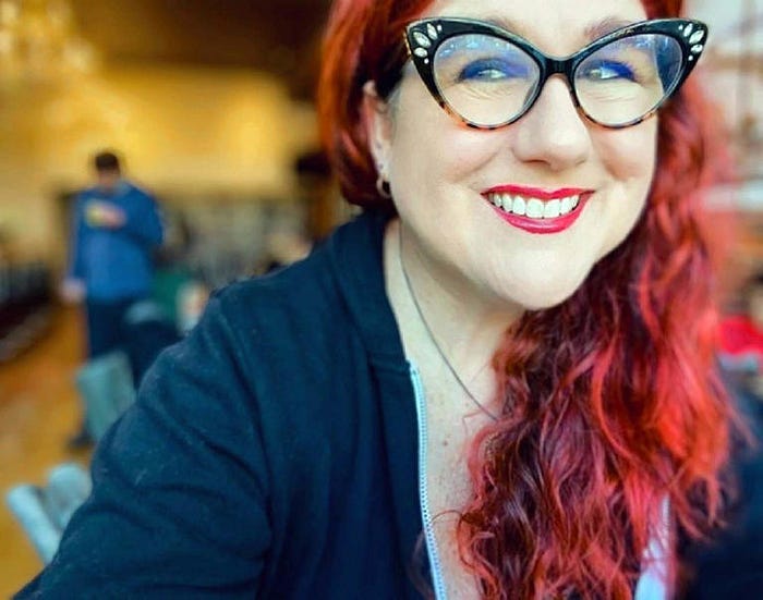 A photo of the author at a cafe. She has red hair and lipstick, cat-eye glasses, and is wearing a hoodie.