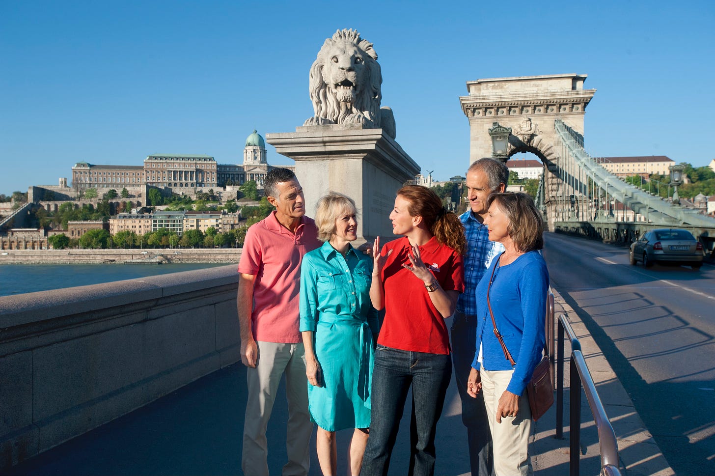 A group of people and a guide stand on a stone bridge in Budapest, with a lion statue and the Chain Bridge visible A group of people and a guide stand on a stone bridge in Budapest, with a lion statue and the Chain Bridge visible