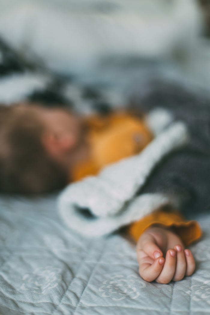 Hazy image of a young child sleeping in bed. The child is sleeping on their back and facing away from the camera. The child has short, dark hair, is wearing a yellow top and has a grey blanket.