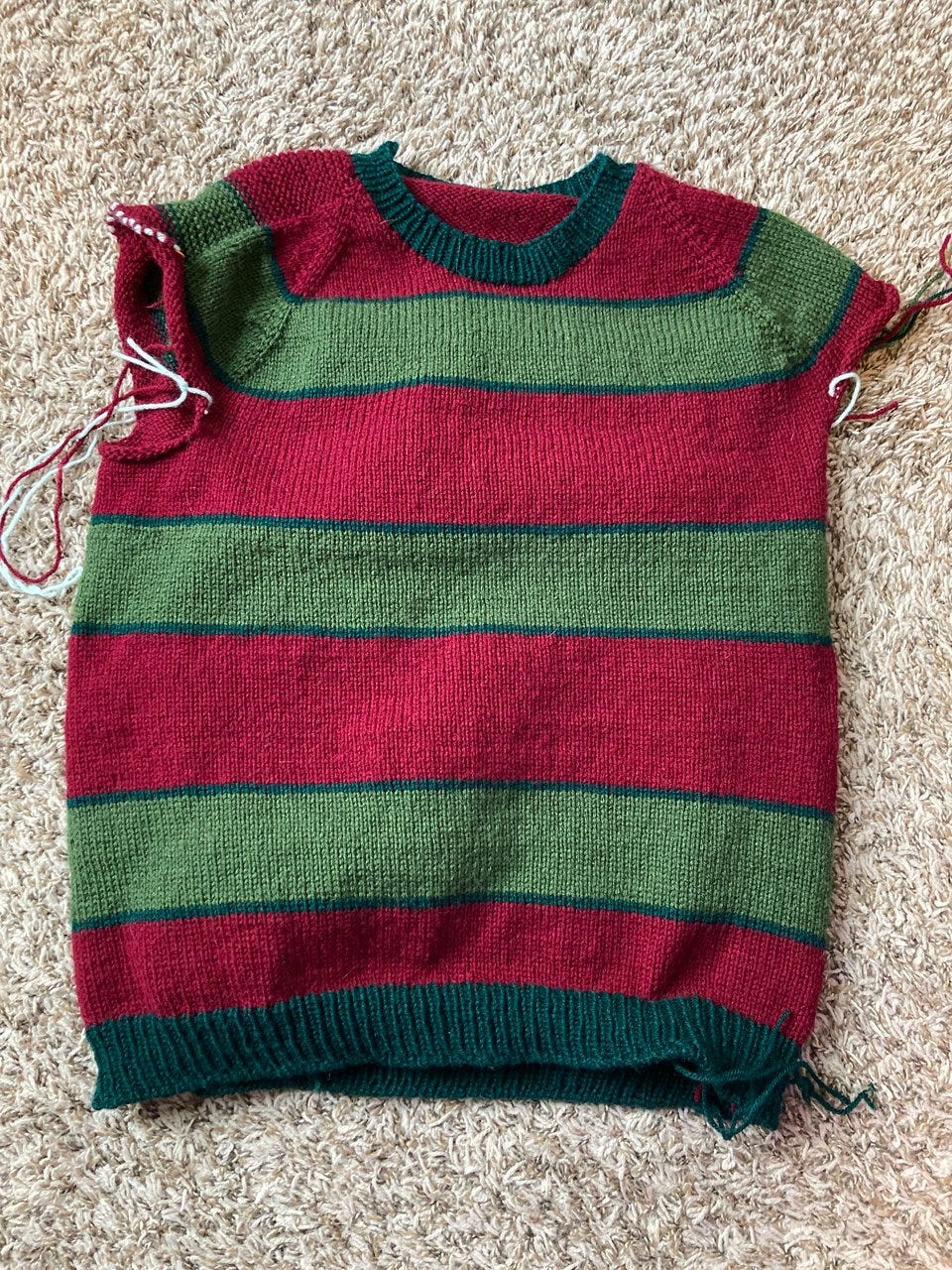A red and green striped sweater on a carpeted floor. The sweater is handknit and missing sleeves/