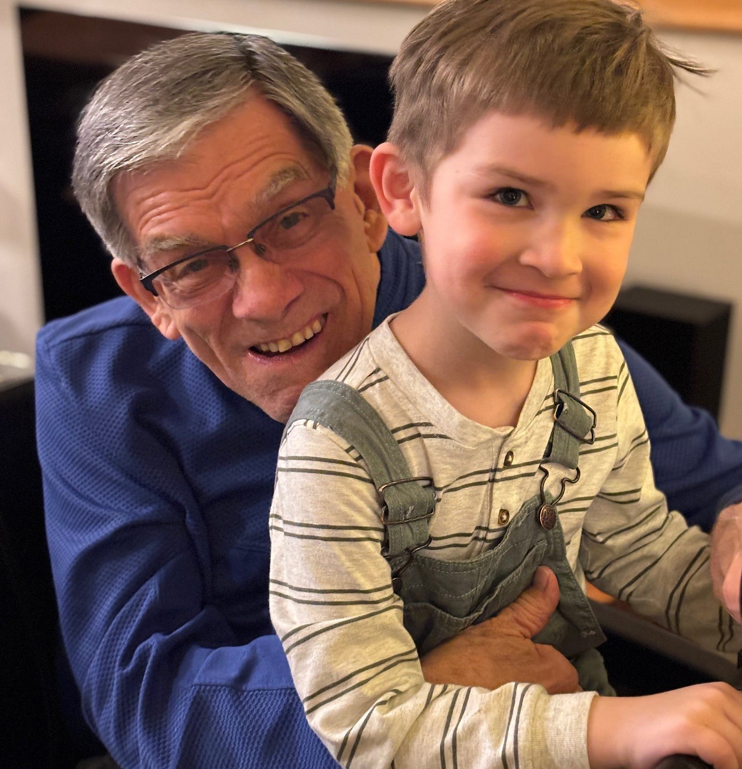 Smiling 82-year-old man with glasses and pepper gray hair snuggles with grinning five-year-old boy in bib overalls.