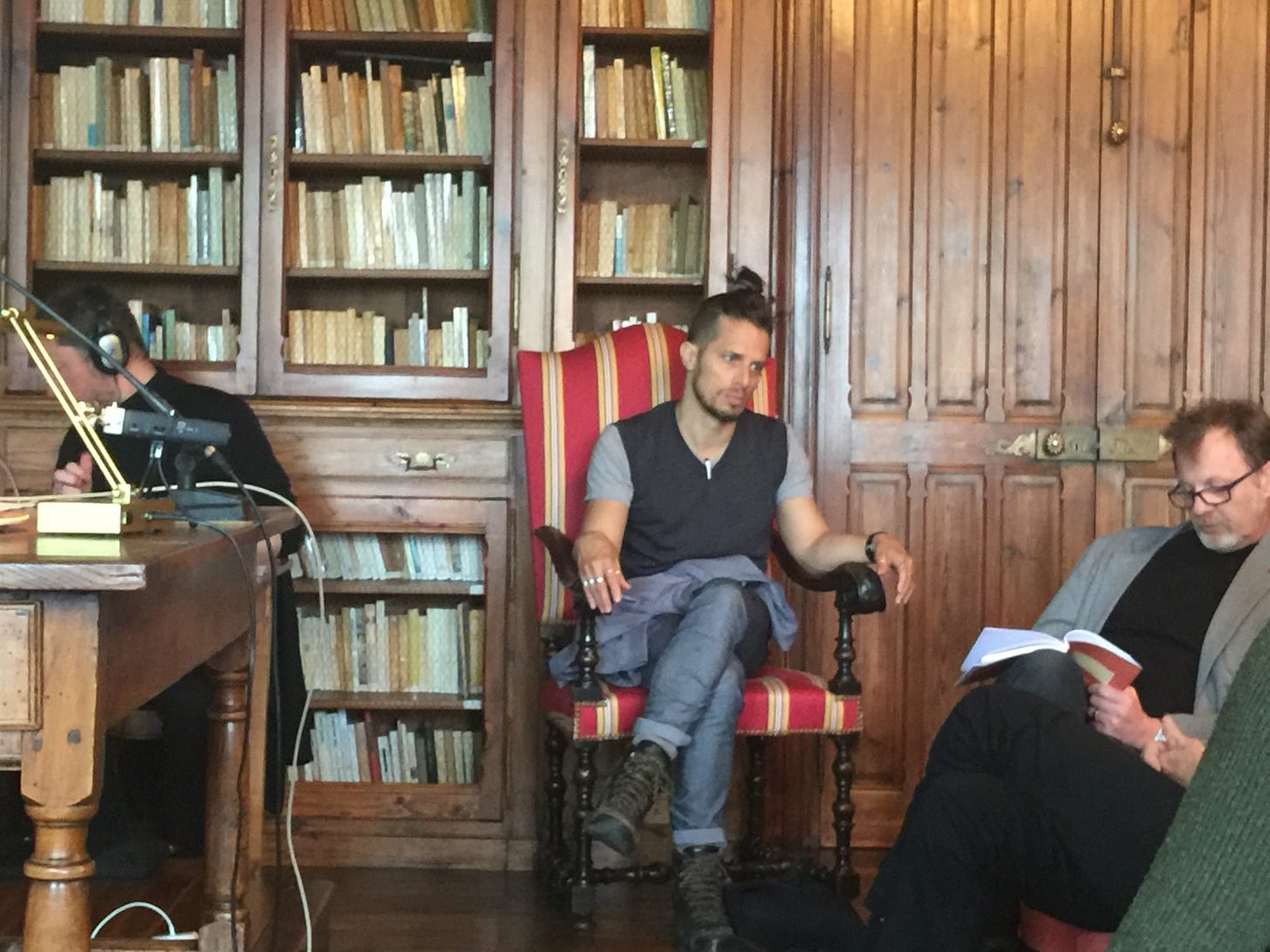 A color photograph of a performance of the Theater of Pure Attention. A man sits, staring to one side in a red and cream striped chair. He is wearing rolled up jeans, hiking boots, a vest over a grey t-shirt. His hair, short on the sides and long on the top, is pulled up into a bun. Behind him are glass-fronted bookshelves filled with books that have pale spines. To the left, behind a desk, is a man with headphones and recording equipment. To the right a bearded man in a jacket is looking down at an open book. The central figure appears preoccupied, or absorbed in thought.