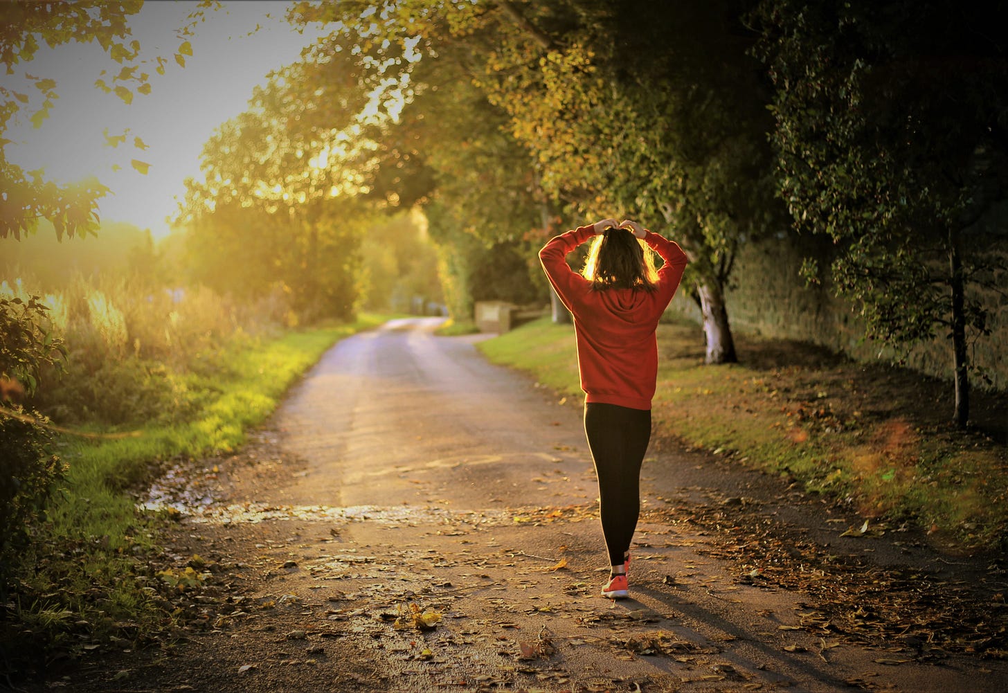 Rear view of woman wearing red shirt and black pants walking on dirt road with hands on top of her head Rear view of woman wearing red shirt and black pants walking on dirt road with hands on top of her head