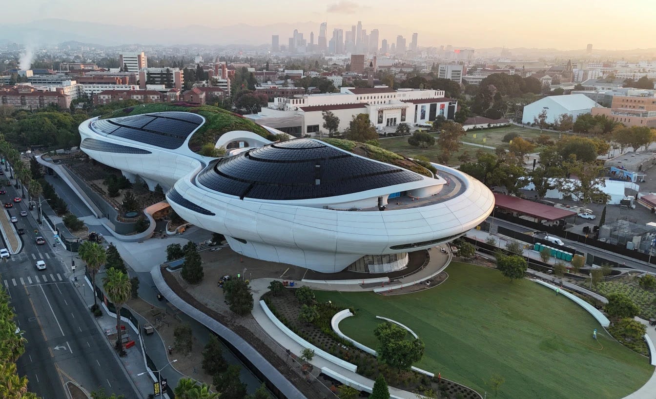 A white building in an infinity shape with black, glass roofing. Off to the left is a street with a few cars driving by. In front the white building is a large grass area. A white building in an infinity shape with black, glass roofing. Off to the left is a street with a few cars driving by. In front the white building is a large grass area.