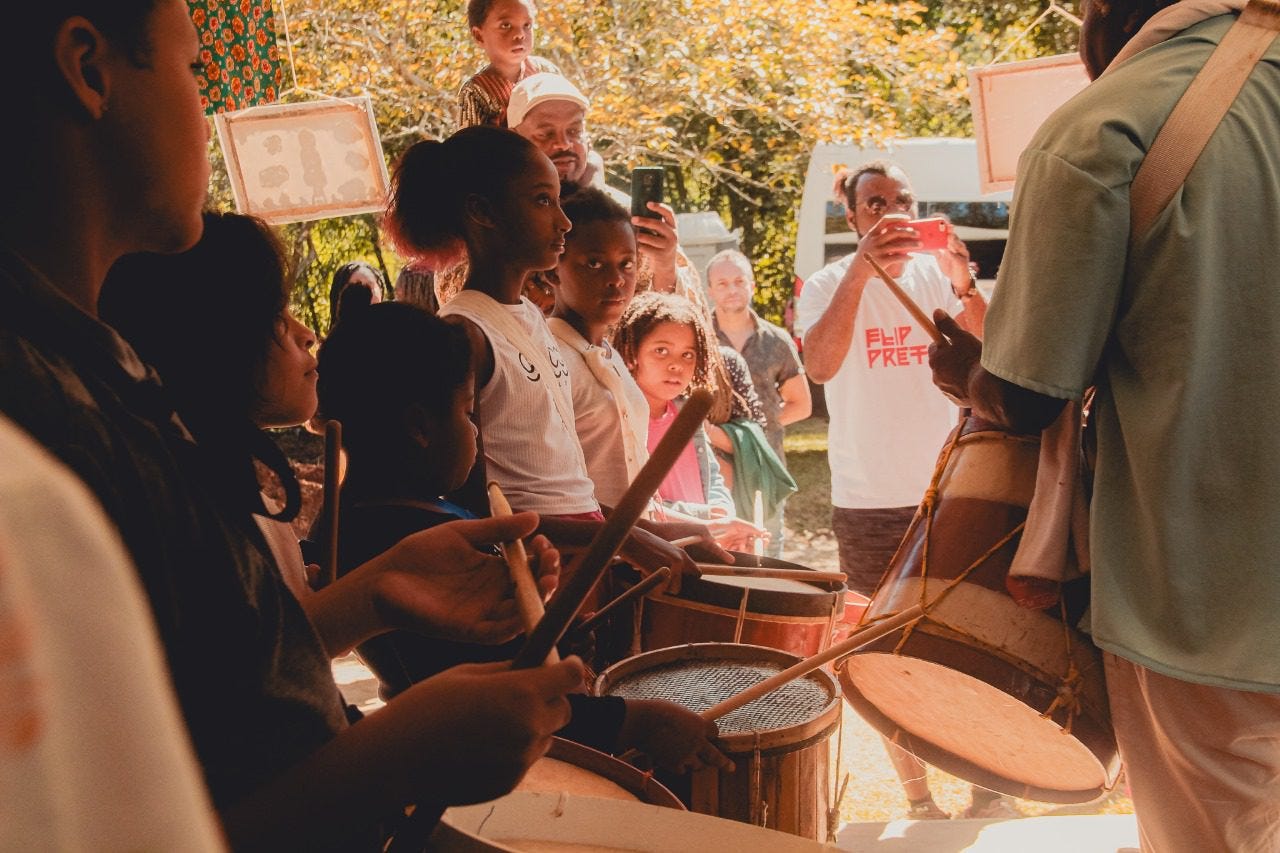 #descrevemospravocê  A imagem mostra uma oficina de percussão ao ar livre, com a participação de crianças e a presença de adultos observando. Em primeiro plano, do lado esquerdo, um grupo de crianças toca tambores com baquetas. Elas estão alinhadas lado a lado, concentradas, com os instrumentos apoiados à frente. À direita, um homem de costas, vestido com camisa verde-clara, segura um grande tambor pendurado por uma alça no ombro, conduzindo a atividade.  Ao fundo, adultos acompanham a cena com interesse. Um homem usa uma camiseta branca com a palavra “FLIP PRETA” estampada em vermelho e grava o momento com o celular. Atrás dele, outras pessoas observam e sorriem. Uma criança pequena está sentada sobre os ombros de um adulto, olhando atenta para a atividade. O ambiente é ensolarado, com árvores de folhas amarelas e verdes ao fundo, sugerindo um clima agradável de outono ou primavera. Cortinas estampadas e molduras penduradas compõem a decoração do espaço.  A atmosfera é alegre, de aprendizado coletivo e celebração cultural, com destaque para a valorização da cultura afro-brasileira e a participação de crianças negras.