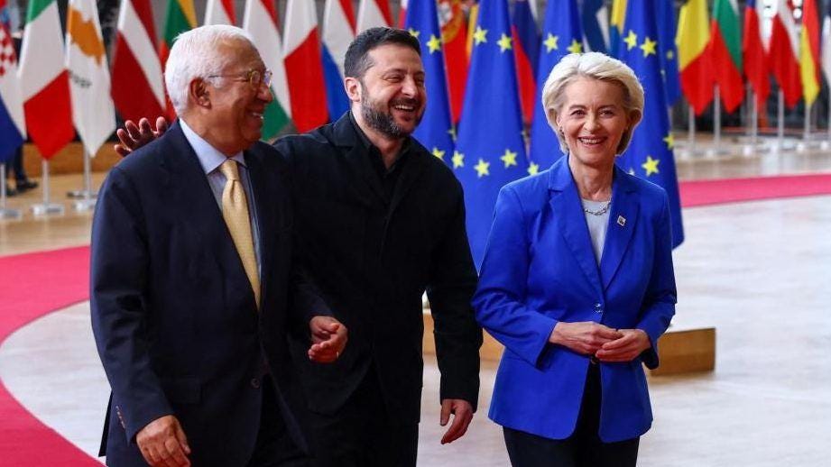 António Costa, Volodymyr Zelensky and Ursula Von der Leyen laughing as they walk, with many country flags hanging behind them. António Costa, Volodymyr Zelensky and Ursula Von der Leyen laughing as they walk, with many country flags hanging behind them.