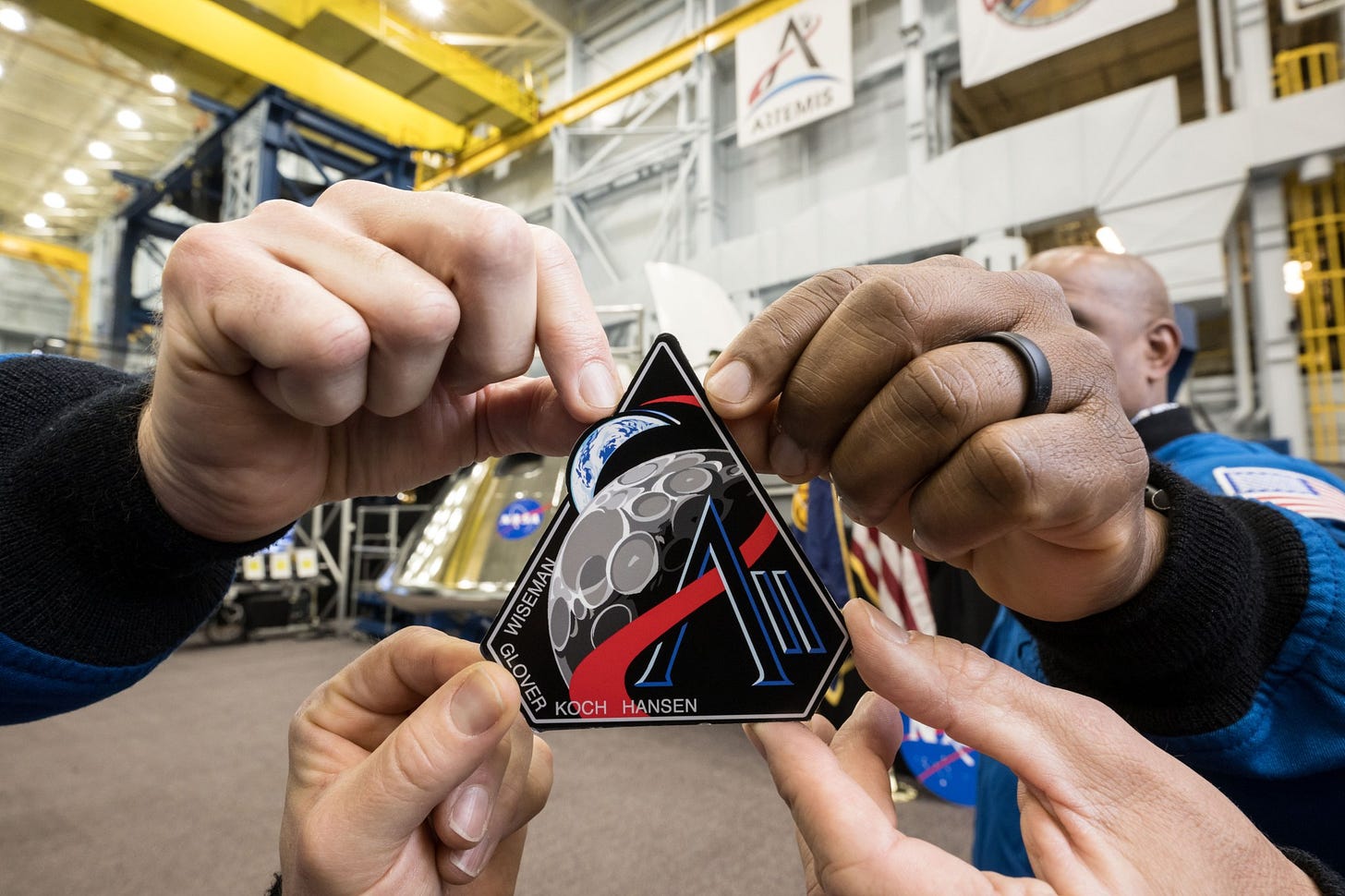 Four hands hold onto a corner of the Artemis II mission patch. The patch is five-sided, with a black background. The last names of the crew – Wiseman, Glover, Koch, and Hansen – are written along the patch’s bottom left corner. The Earth and Moon are visible on the patch, reminiscent of the famous “Earthrise” photo. A red line curves through the lines of a letter “A,” then around the Moon and Earth. Next to the “A” are two lines that look like the Roman numeral “I”: this represents Artemis II. Four hands hold onto a corner of the Artemis II mission patch. The patch is five-sided, with a black background. The last names of the crew – Wiseman, Glover, Koch, and Hansen – are written along the patch’s bottom left corner. The Earth and Moon are visible on the patch, reminiscent of the famous “Earthrise” photo. A red line curves through the lines of a letter “A,” then around the Moon and Earth. Next to the “A” are two lines that look like the Roman numeral “I”: this represents Artemis II.