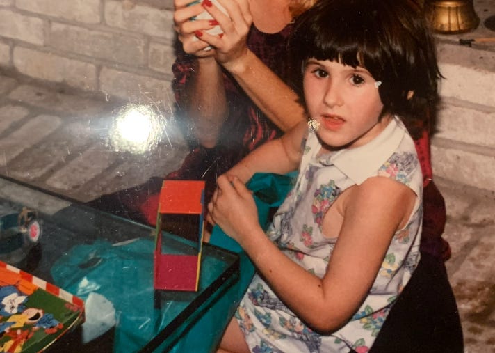 Young Lyric, caught off guard by a camera flash, sits at a glass table next to a person with red fingernails. They seem confused.