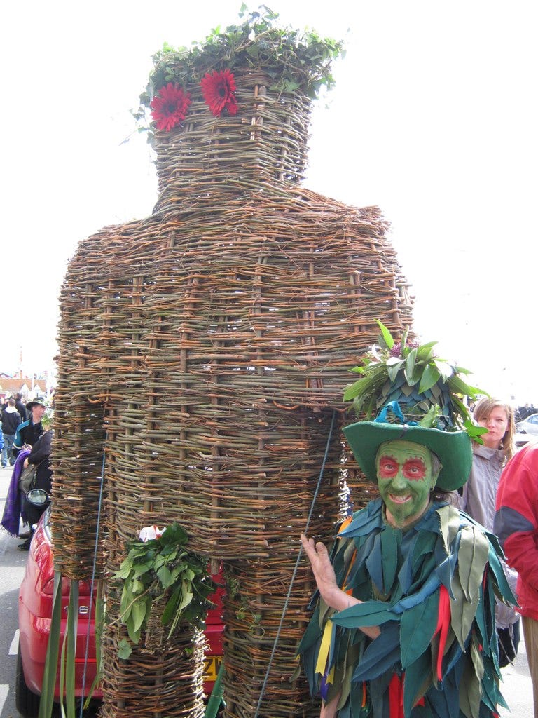 An overexposed photo of a wickerman with flower eyes and ivy hair standing next to a man in green rags, gesturing to it as if introducing a friend