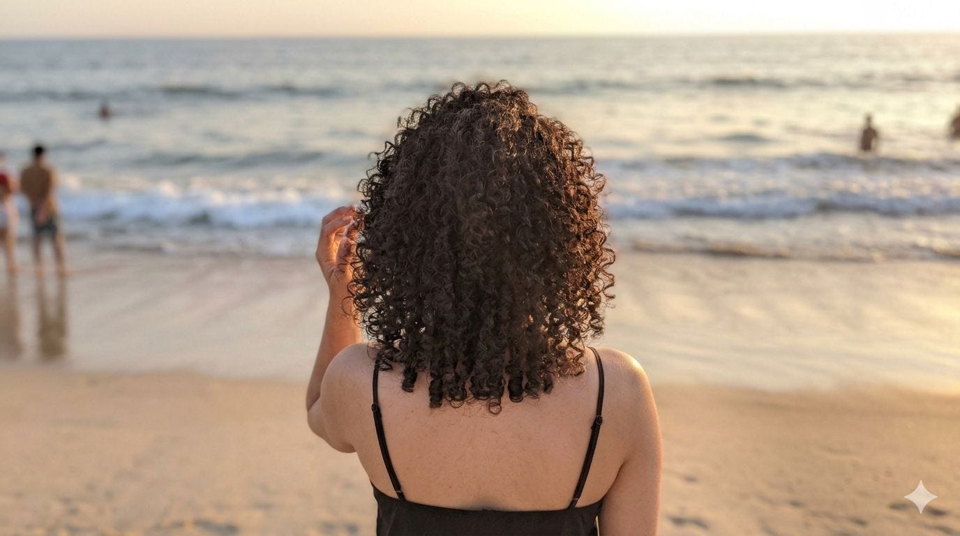 A woman with neck-length dark curly hair and olive skin stands on the beach at sunset looking out at the sea