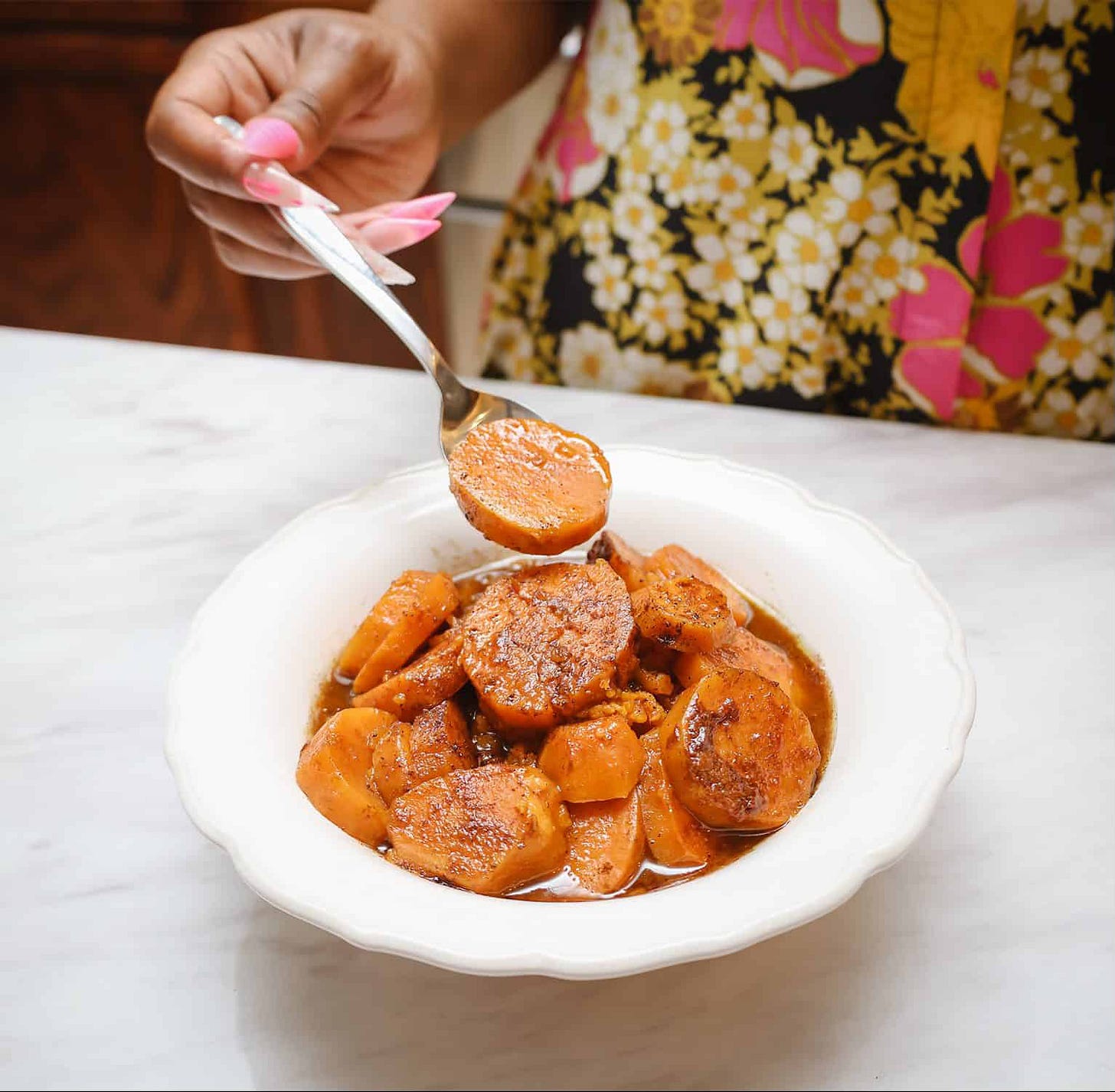 Southern soul food candied yams in a white bowl, topped with buttery brown sugar glaze. Soul food expert and author, Shaunda Necole lifts a spoonful to show the tender, sweet potato rounds — a classic Black folks’ Thanksgiving side dish.