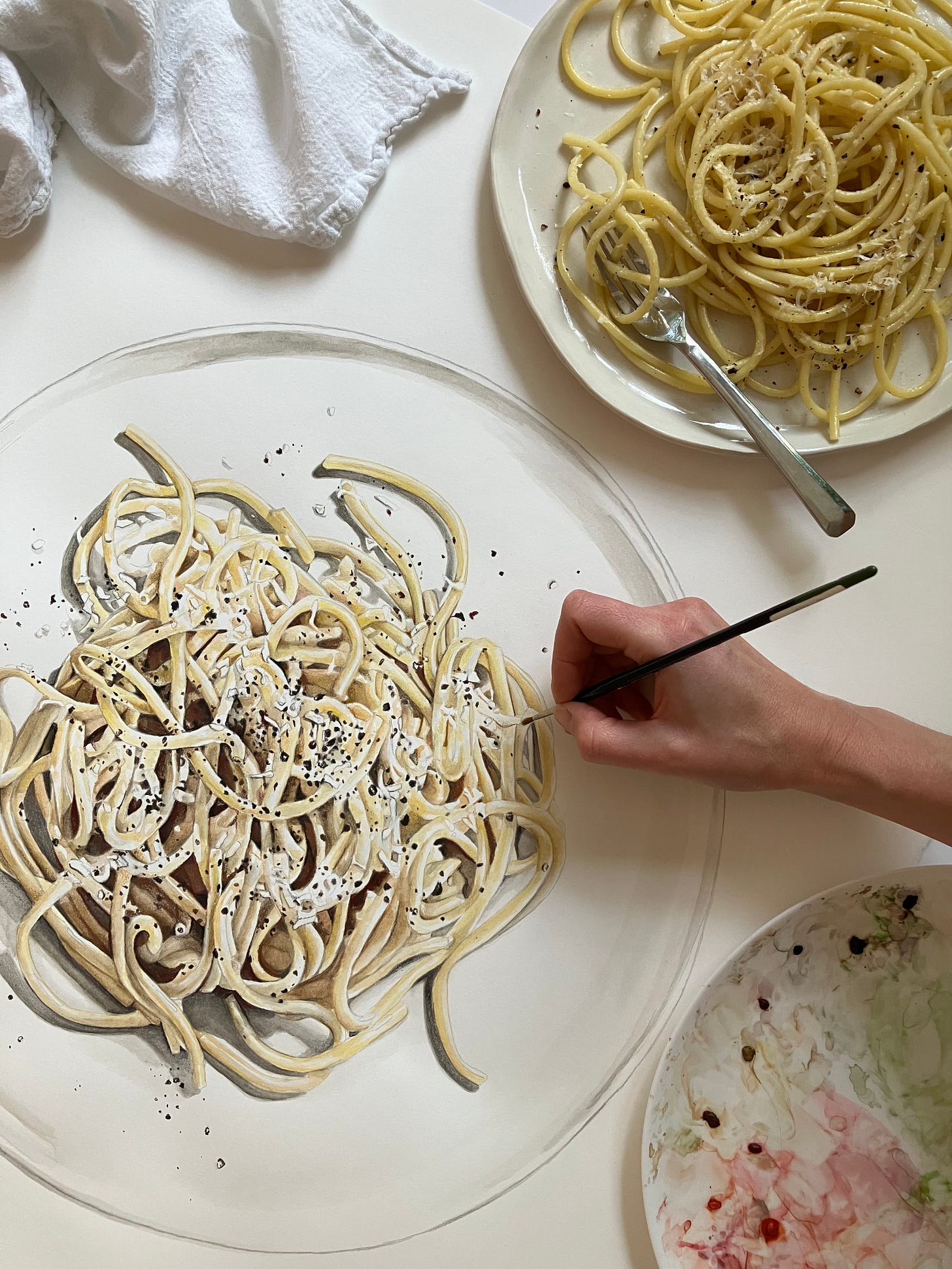 shot of painting from above with plate of pasta sitting to the top right corner. Paints in the bottom right corner and hand with paintbrush over the painting. 