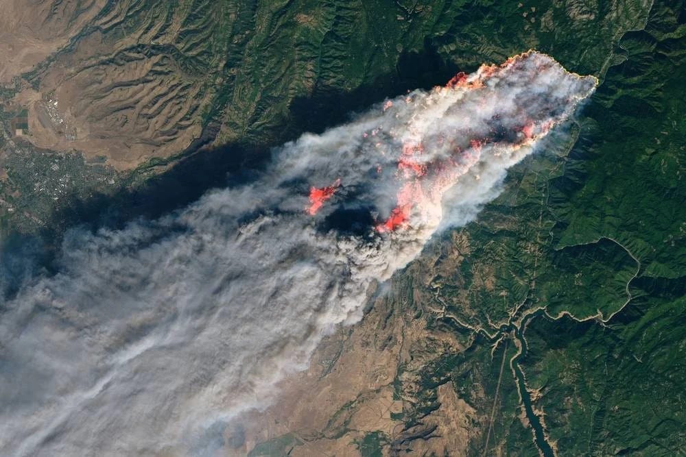 Aerial satellite image of a large wildfire with bright orange flames and extensive smoke plume spreading across forested terrain.