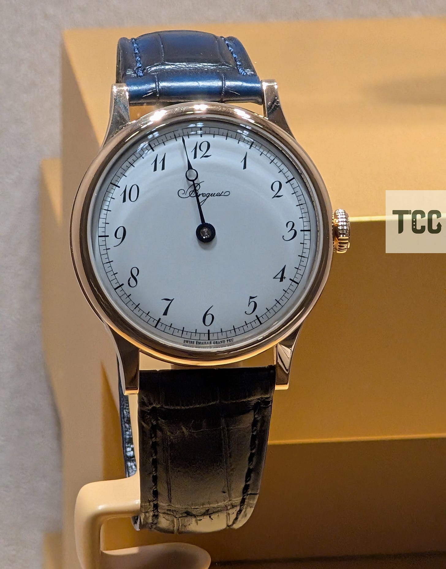 Close-up of a Breguet wristwatch with a white enamel dial, Arabic numerals, and a blue leather strap displayed on a creme stand.
