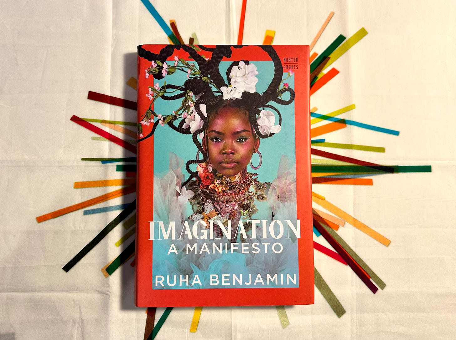 Picture of a book titled “Imagination: A Manifesto” by Ruha Benjamin on a white tablecloth with multi coloured block stripes emanating in different directions from the centre so it looks likethe book is in the middle of a rainbow coloured explosion. The cover of the book features a young Black girl with elaborate braids staring out head-on with a dark orange border and sky-blue background. Picture of a book titled “Imagination: A Manifesto” by Ruha Benjamin on a white tablecloth with multi coloured block stripes emanating in different directions from the centre so it looks likethe book is in the middle of a rainbow coloured explosion. The cover of the book features a young Black girl with elaborate braids staring out head-on with a dark orange border and sky-blue background.