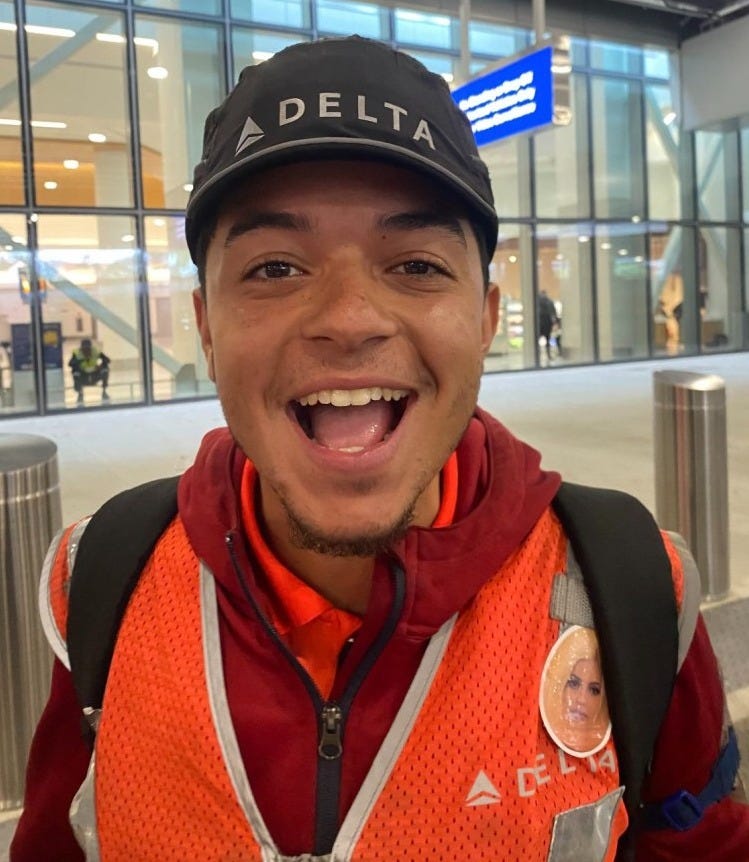 Pena in a Delta cap and a bright orange delta vest, smiling at the camera while at the airport Pena in a Delta cap and a bright orange delta vest, smiling at the camera while at the airport