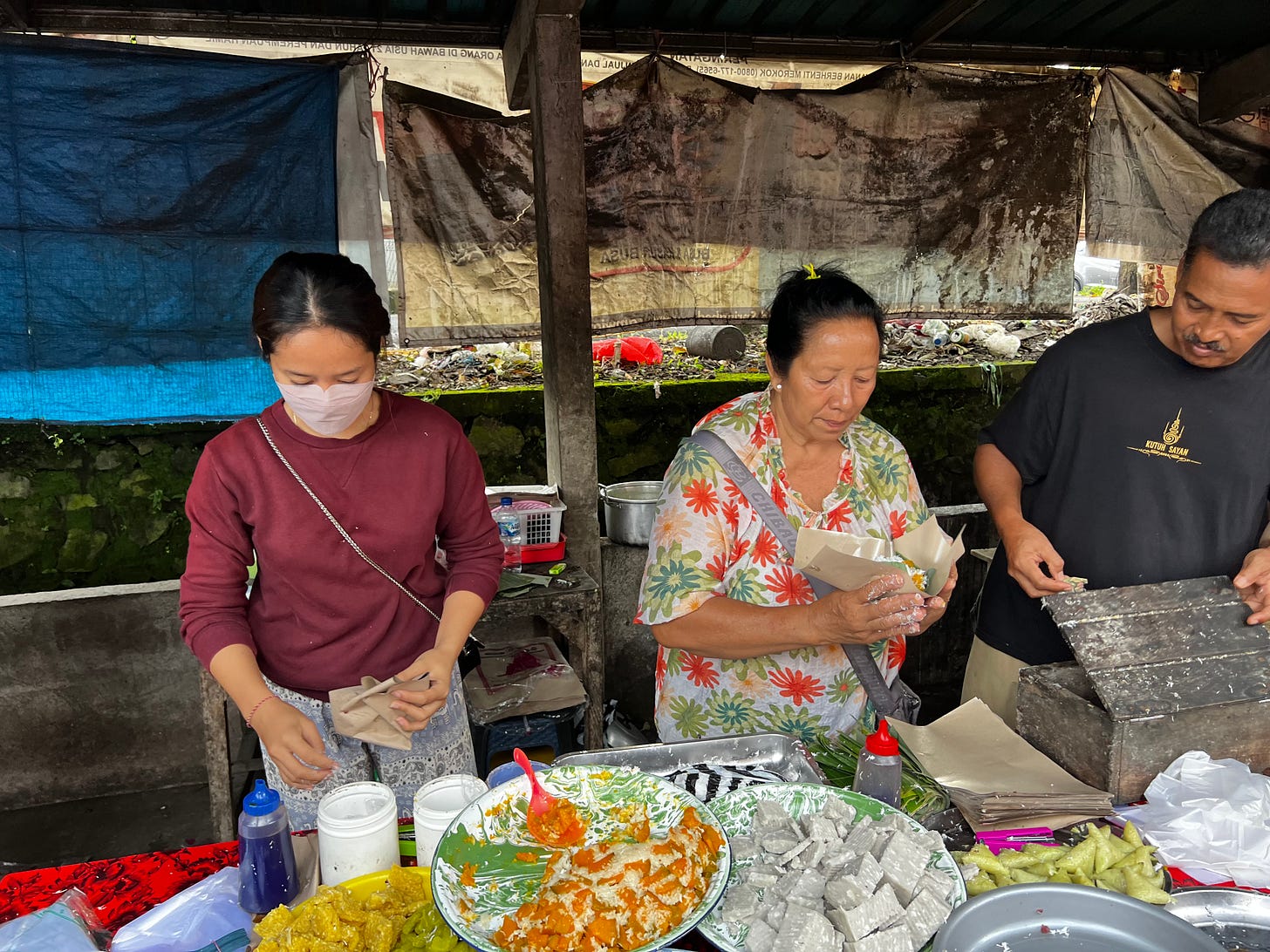 Vendors at a market stall preparing and wrapping food, enamel platters of brightly colored sweets in front of them. 