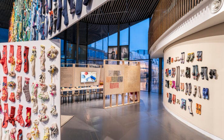Photo of an art exhibition with multicoloured socks with visible darned holes hanging from white walls in a circular building Photo of an art exhibition with multicoloured socks with visible darned holes hanging from white walls in a circular building