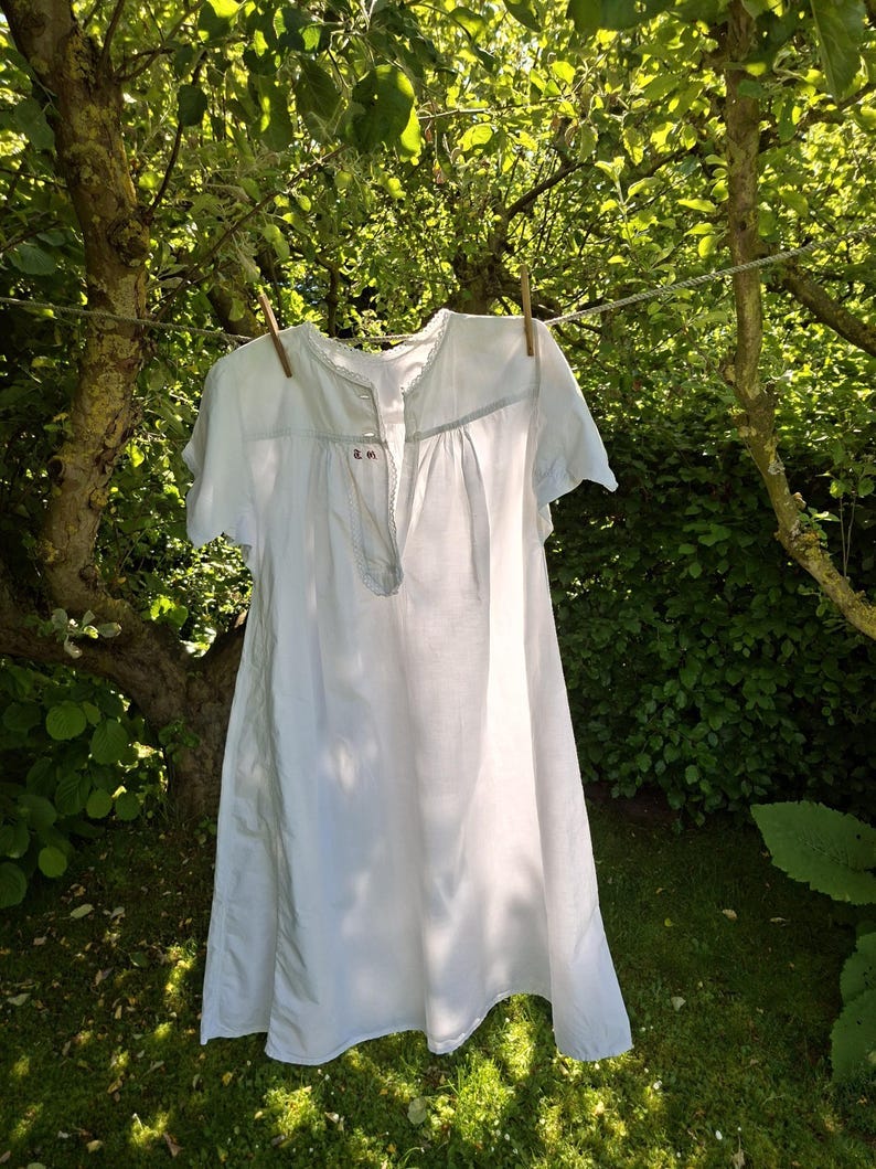 May include: A white, short-sleeved garment, possibly a nightgown or dress, hangs on a clothesline with wooden clothespins. The garment is made of a light fabric and has a button-up front. The background features lush green foliage and sunlight.
