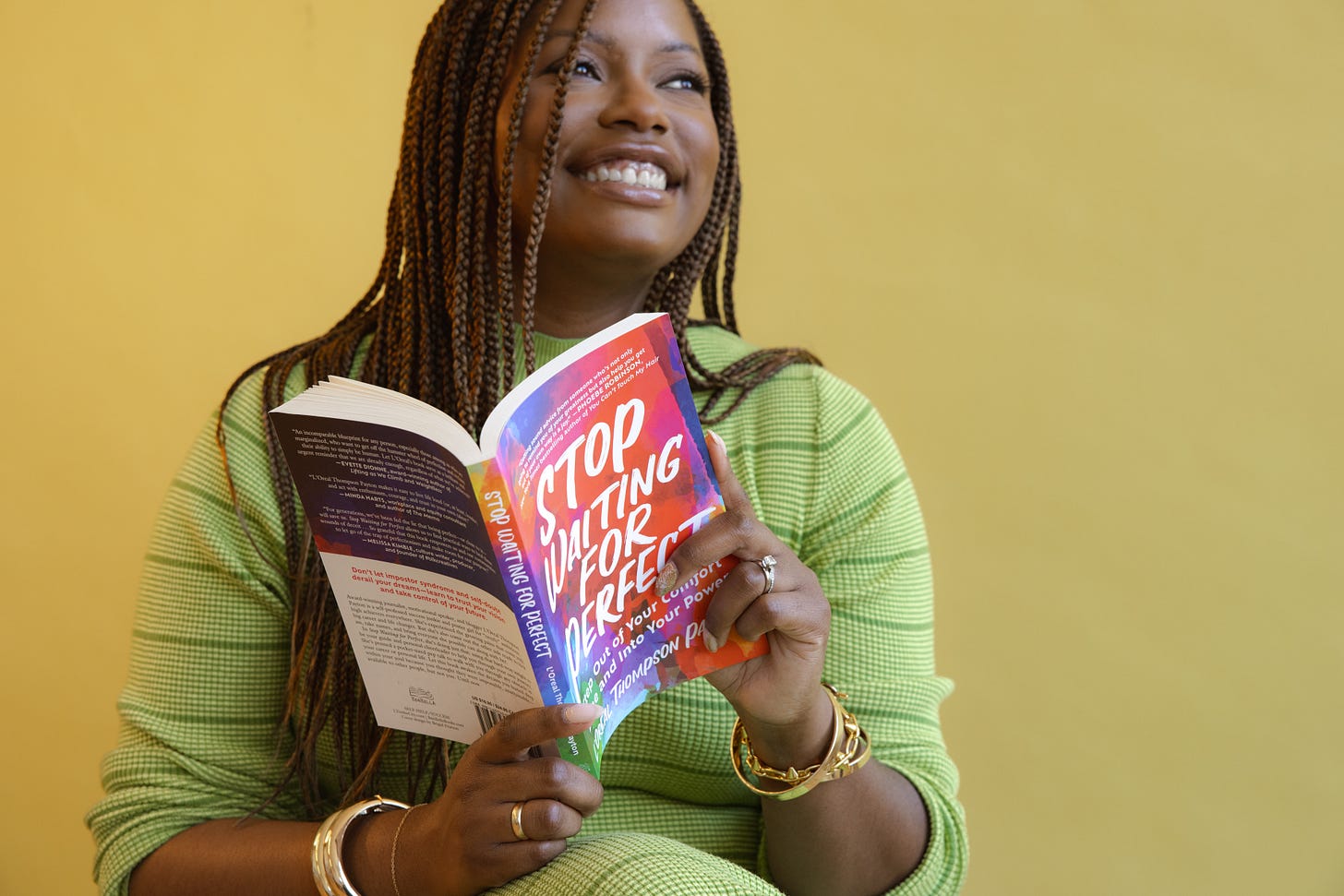 Picture of a Black woman with braids wearing a green dress sitting in front of a yellow background holding a book