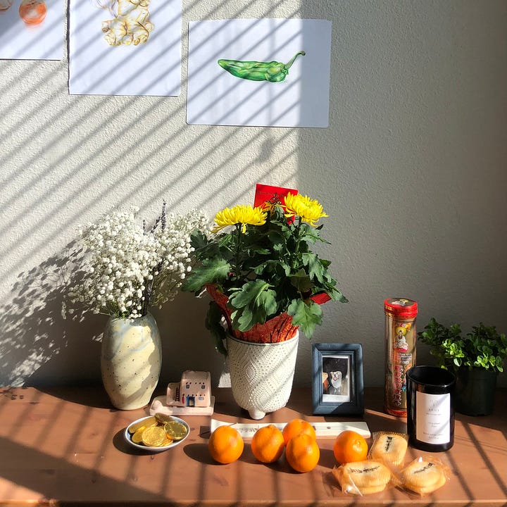 On the left is an ancestral shrine with white and yellow flowers, a framed photo of Jenn's dog Coco, a tube of incense stickers, clementines, a tray of gold chocolate coins and rice crackers. On the right is a tablespread of food, including dumplings, tteokbokki, rice and salad.