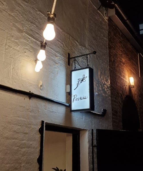 A white painted brick building with bulb fairy lights around it and a neon sign that says Piccalilli