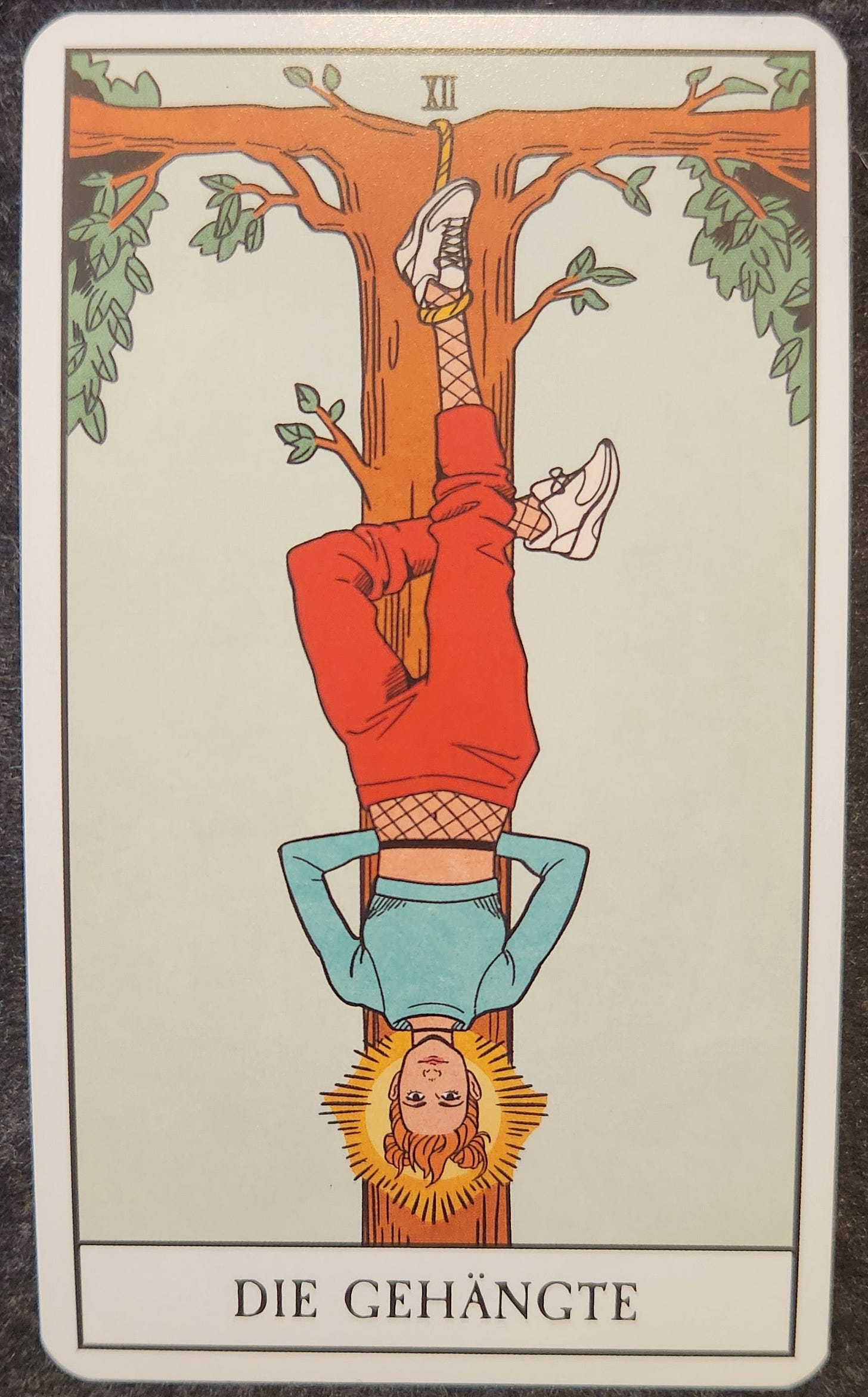 A tarot card labelled XII Die Gehängte. A person is hanging upside down on a tree by one leg. Her other leg is bent and crossed behind the hanging leg. Her arms are crossed behind her back. Her face is neutral. There is a halo of golden light around her head. 