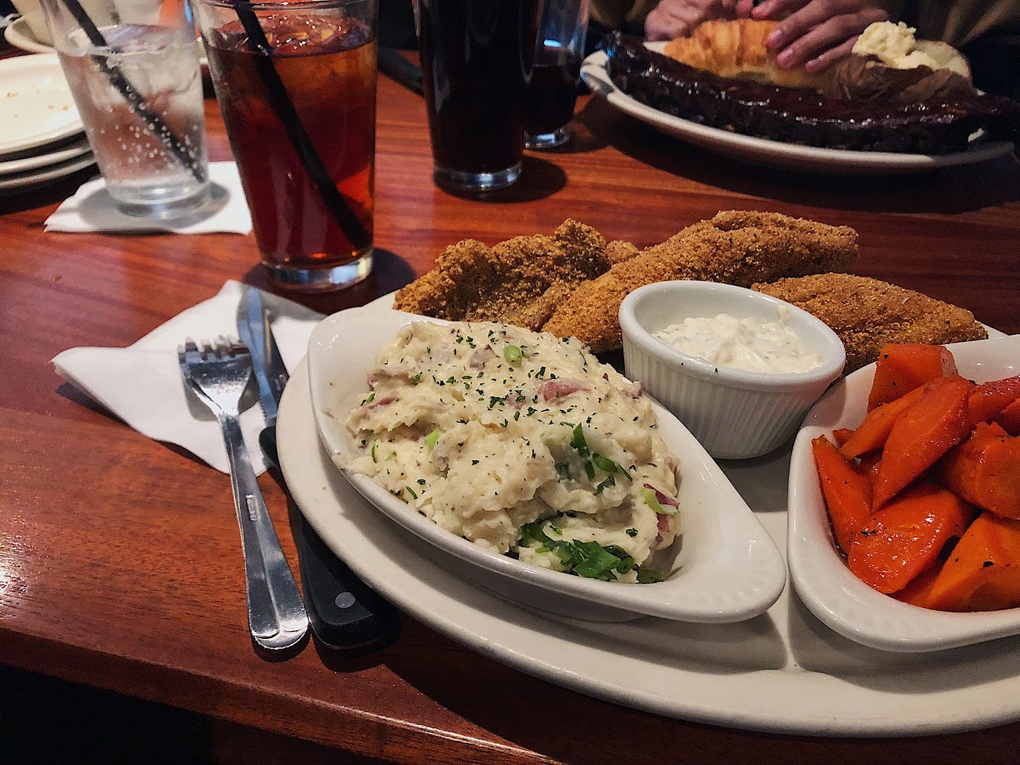 A plate of fried catfish, mashed potatoes, and glazed carrots on a wooden table with silverware, and three drinks in glassed in frame