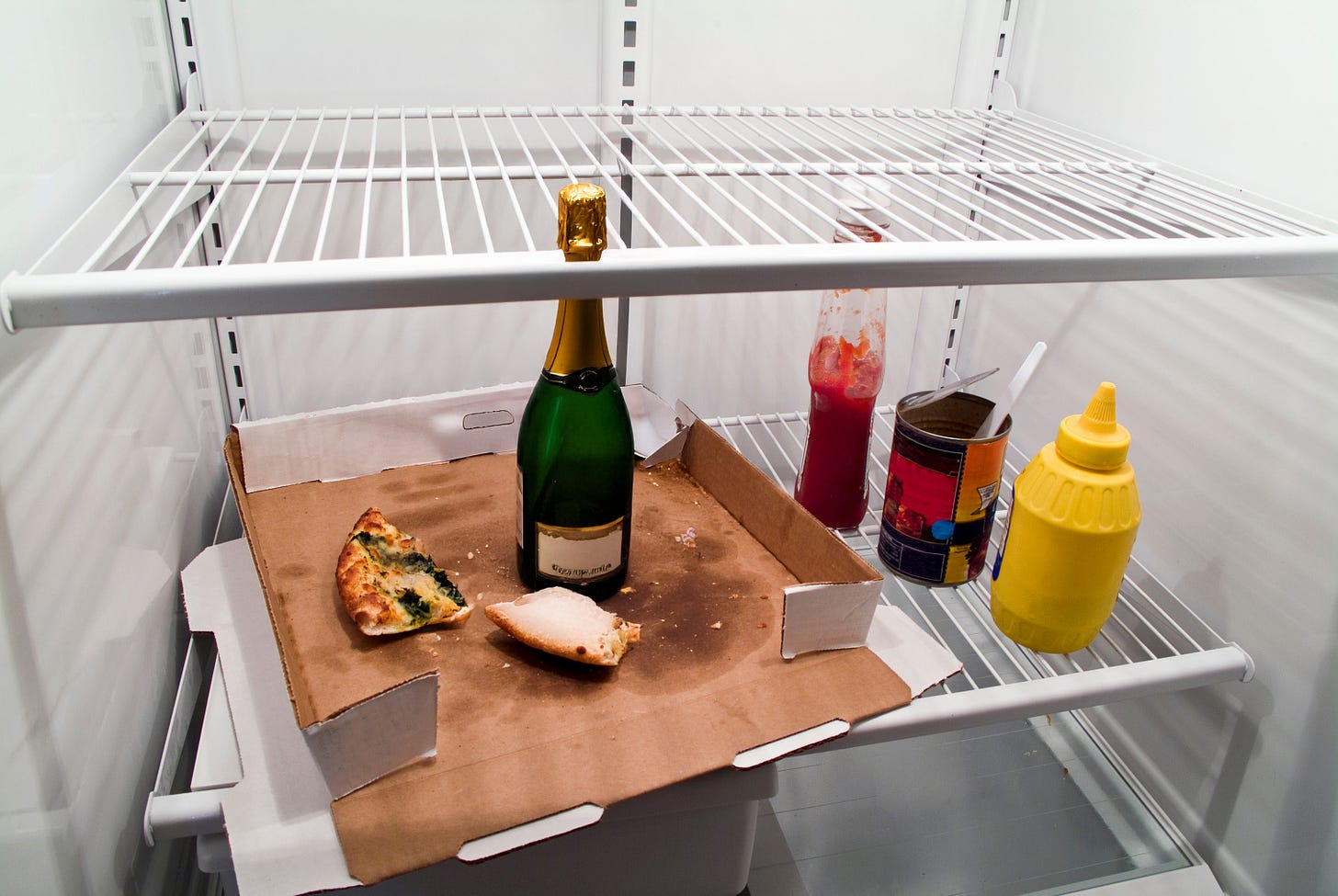 Mostly empty refrigerator with a pizza box inside containing a few slices of pizza; next to it our some condiments and a bottle of champagne (a new addition) Mostly empty refrigerator with a pizza box inside containing a few slices of pizza; next to it our some condiments and a bottle of champagne (a new addition)