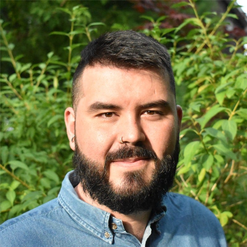 Headshot photo of a smiling Yahya, with a full beard, short hair, and a blue collared shirt, in front of green plants.