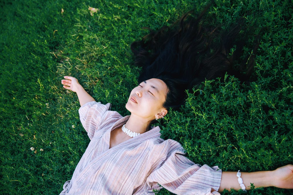 A photo of an Asian person with long black hair, their eyes closed, lying in a field with their arms outstretched. A photo of an Asian person with long black hair, their eyes closed, lying in a field with their arms outstretched.