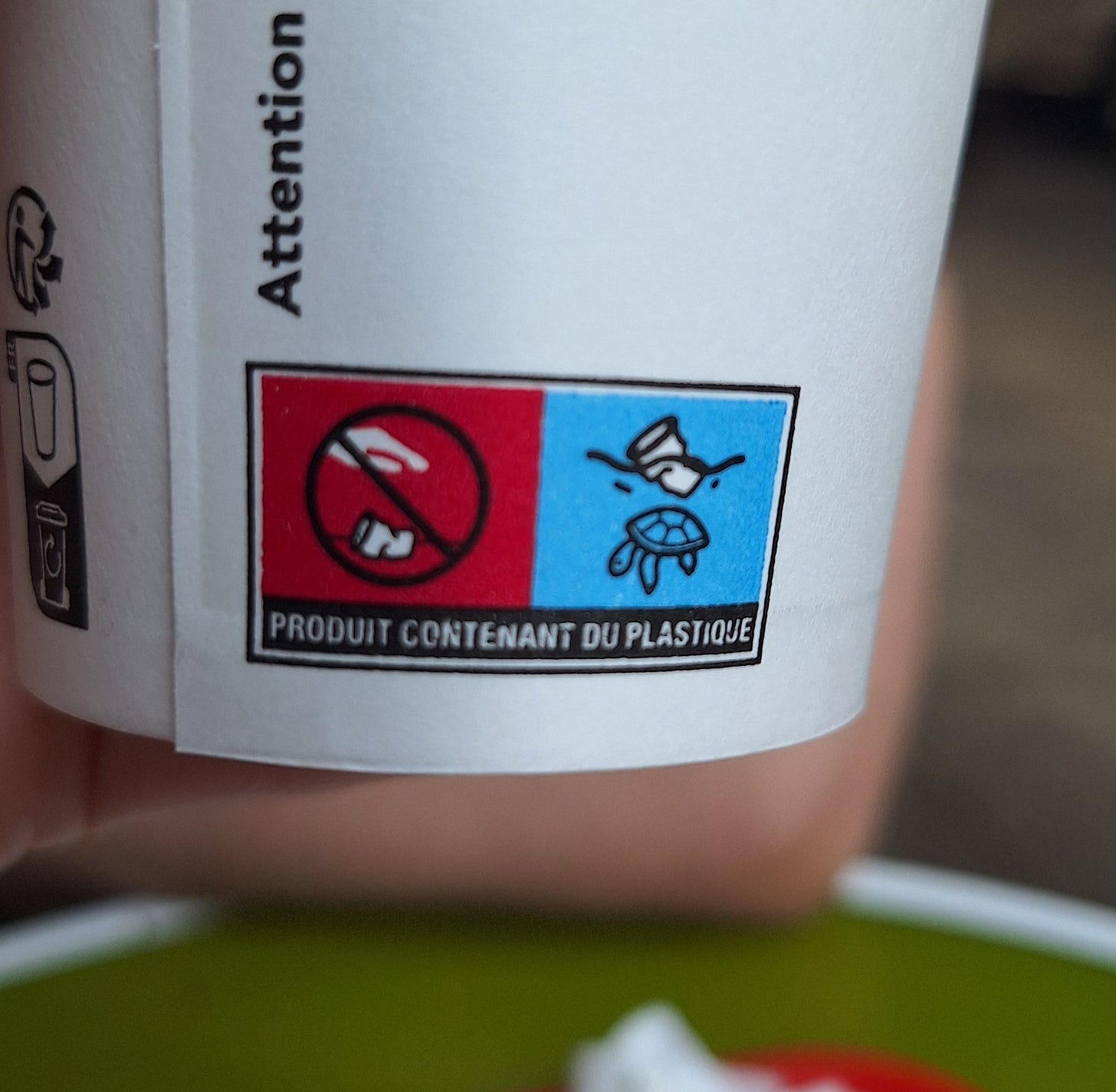 Foto. Ícones num copo descartável: Num fundo vermelho tem um copo amassado no chã, uma mão acima dele e está com um símbolo de proibido, num fundo azul tem um copo boiando na superfície da água e uma tartaruga nada sob ele. Embaixo o texto "produit contenant du plastique"