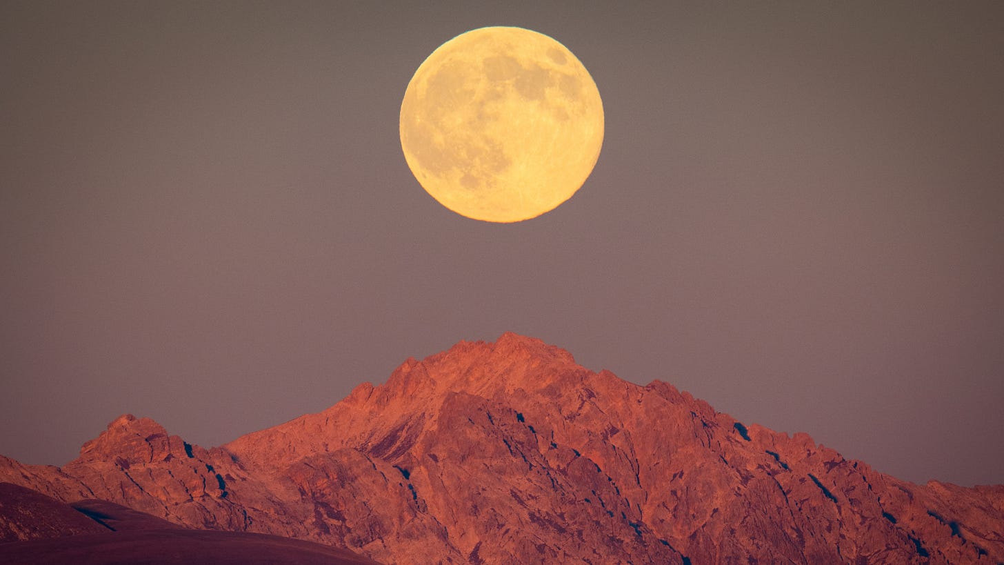 A yellow full moon is pictured rising above a rocky mountain against a pale grey-blue sky.