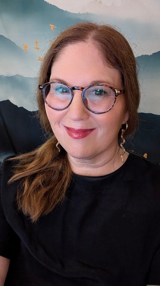 Portrait accompanying the Jenna Matlin Tarot DMs interview, showing a smiling woman with auburn hair, round glasses and a dangling snake-shaped earring against a misty mountain backdrop. Portrait accompanying the Jenna Matlin Tarot DMs interview, showing a smiling woman with auburn hair, round glasses and a dangling snake-shaped earring against a misty mountain backdrop.