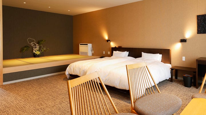 Image 1 alt text: An accessible bathroom with a natural stone soaking tub, grab bars, handheld shower, and a pink mechanized shower chair lift positioned beside the bath for wheelchair transfers.  Image 2 alt text: A spacious Japanese-Western style guest room with twin beds, warm lighting, wood furniture, and a raised tatami platform decorated with a floral arrangement.