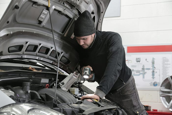 A man looking inside the car hood with a flashlight trying to diagnose what’s happening. A man looking inside the car hood with a flashlight trying to diagnose what’s happening.