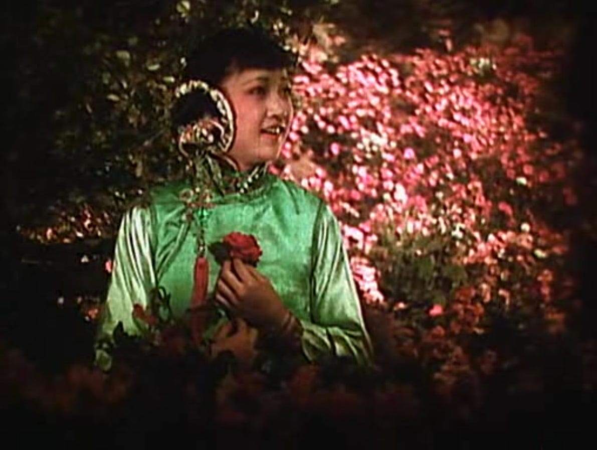 a young Chinese woman in a traditional green dress, grasps a bunch of flowers, standing in front of a backdrop of vibrant red/pink flowers