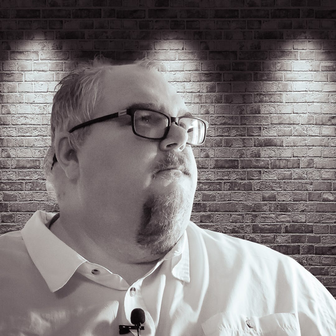 Man in glasses and collared shirt standing in front of a brick wall with spotlight-style lighting looking thoughtfully to the side wearing a clip-on microphone suggesting he is speaking or recording content about mentorship and cultural leadership Man in glasses and collared shirt standing in front of a brick wall with spotlight-style lighting looking thoughtfully to the side wearing a clip-on microphone suggesting he is speaking or recording content about mentorship and cultural leadership
