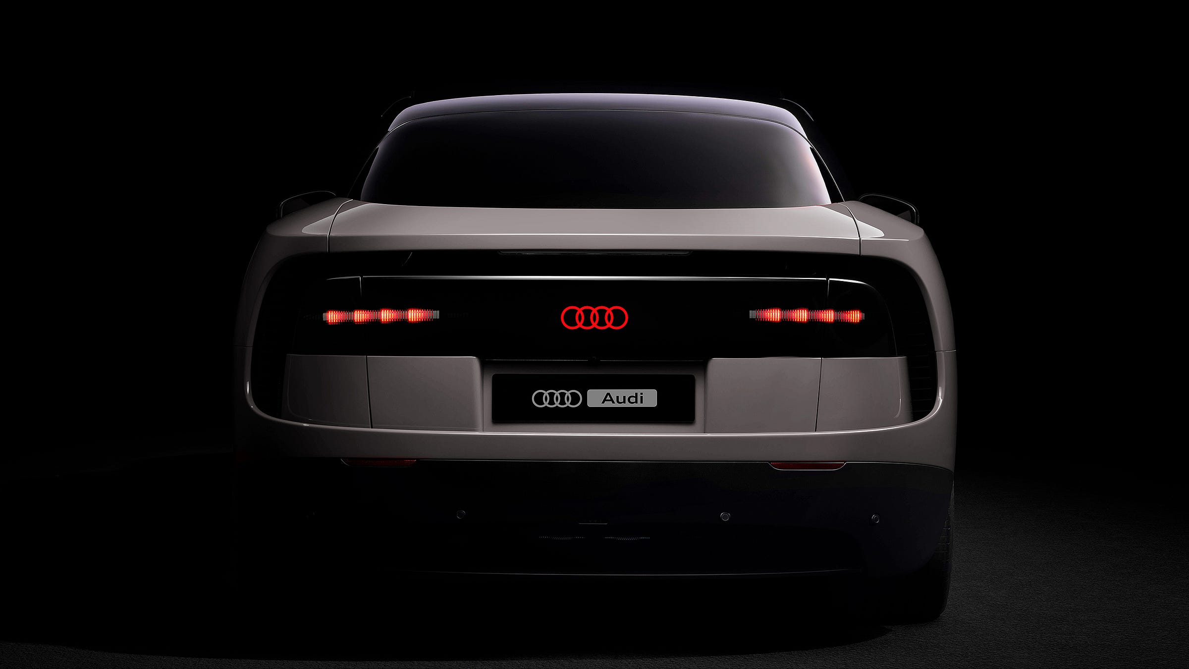 Straight-on rear view of the Ferrari Luce in a dark studio setting, retouched to replace Ferrari branding with Audi's four-ring logo. The car is finished in matte warm grey with a wide black panel spanning the rear, housing two clusters of horizontal LED tail lights flanking a glowing red Audi four-ring emblem at centre. A licence plate below reads "Audi" alongside the four-ring logo.