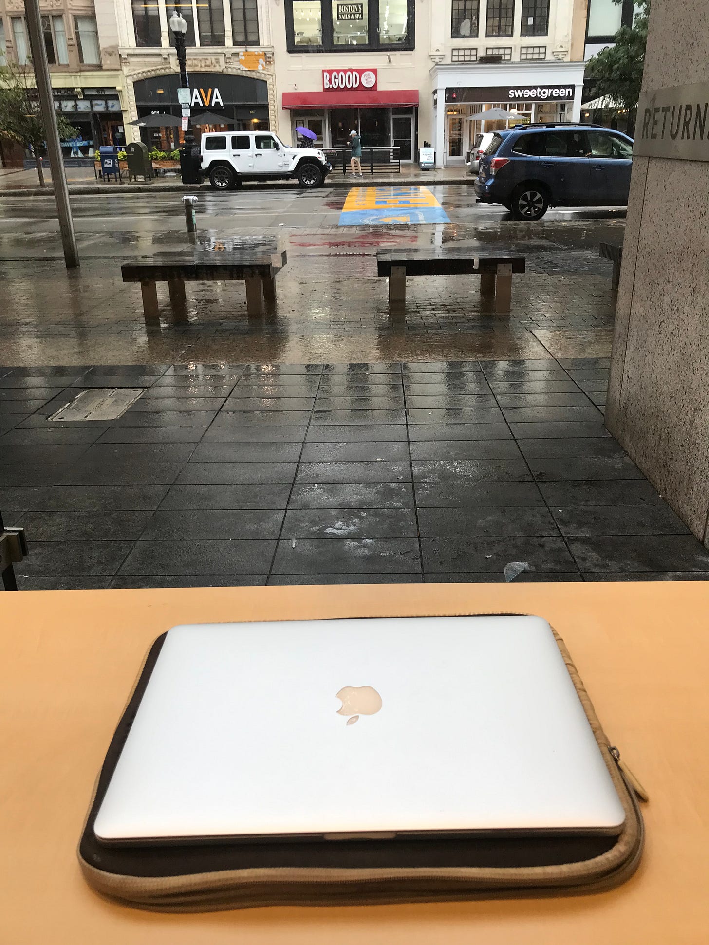 Image of a Macbook laptop closed on a counter beside a big window. The view outside is of a street in Boston, in front of the Boston Public Library, with cars parked and fast-casual restaurants across the street. The Boston Marathon finish line is visible. Image of a Macbook laptop closed on a counter beside a big window. The view outside is of a street in Boston, in front of the Boston Public Library, with cars parked and fast-casual restaurants across the street. The Boston Marathon finish line is visible.
