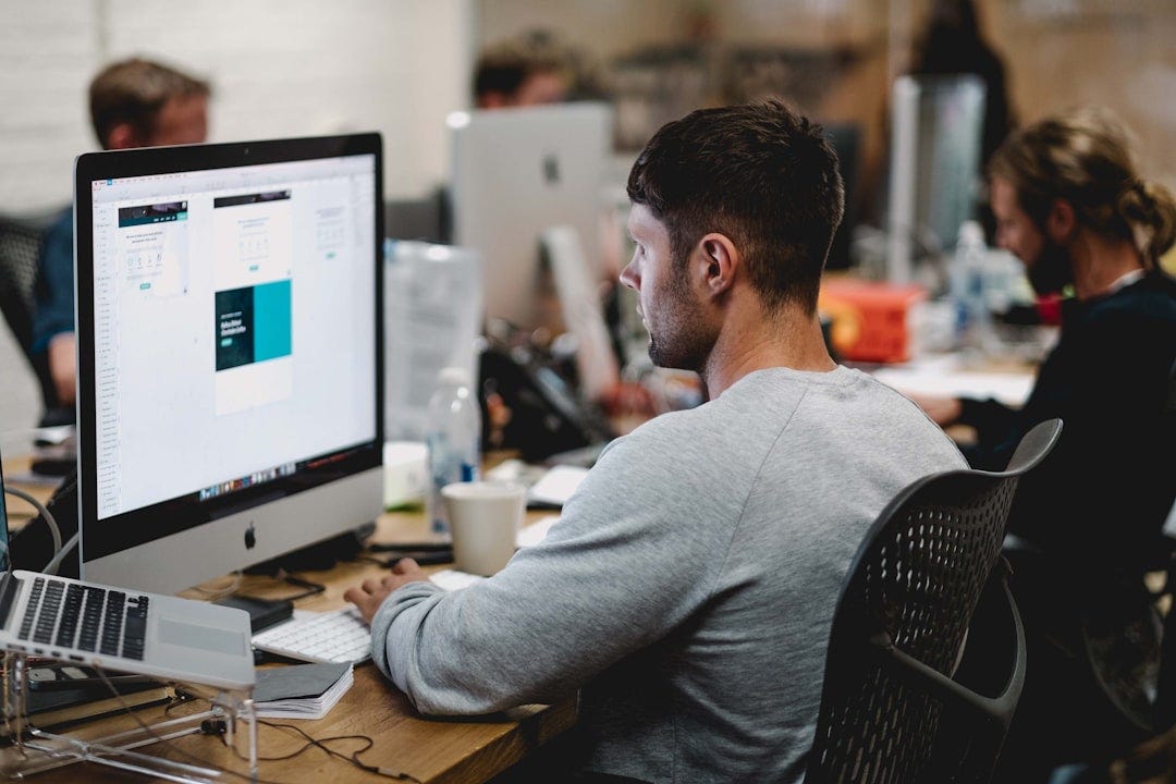 man in gray sweatshirt sitting on chair in front of iMac man in gray sweatshirt sitting on chair in front of iMac