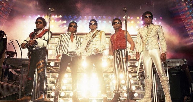Photo of five Jacksons with the most direct inspiration of costuming from the cover illustration on the left with 3 diagonal stripes on one pant leg with the other striped like an upside down T. The jacket is white with red over the shoulders and chest.