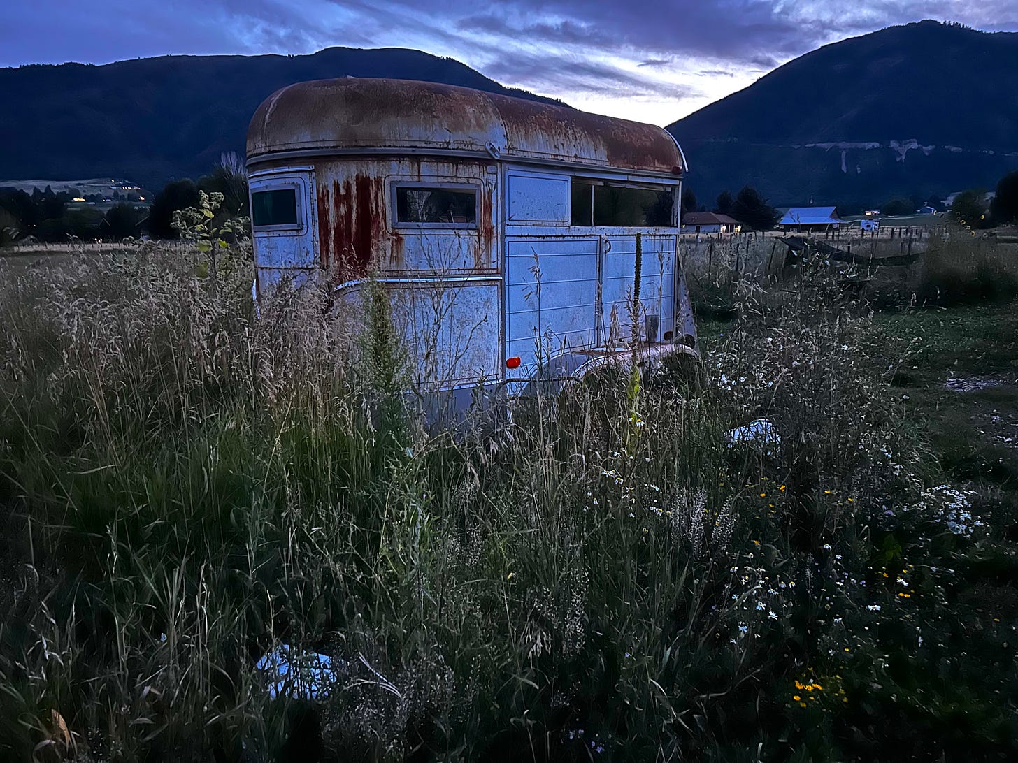 A rusty trailer parked in a field of overgrown grass and wildflowers, its white sides blue in moonlight, in the background two navy-colored peaks between a billowy sky