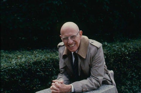 The French philosopher Michel Foucault wears a trench coat and laughs in the Rodin Museum's sculpture garden
