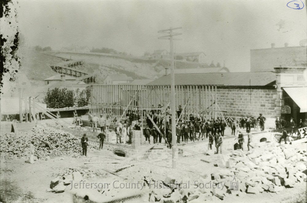 A historic photograph showing workers and early buildings under construction in Port Townsend during the late 1800s.