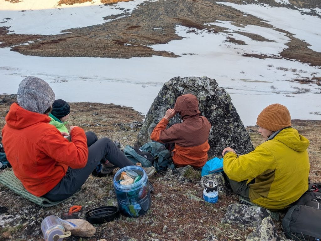 Four people in colorful jackets sit on the tundra eating around a lichen-covered boulder, cooking gear and a bear canister beside them, a snow-patched slope behind in dim evening light.